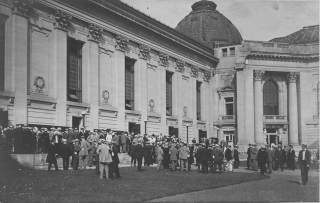 An old, black and white photo of a crowd of alumni in hats and suits in front of a building on the plaza.