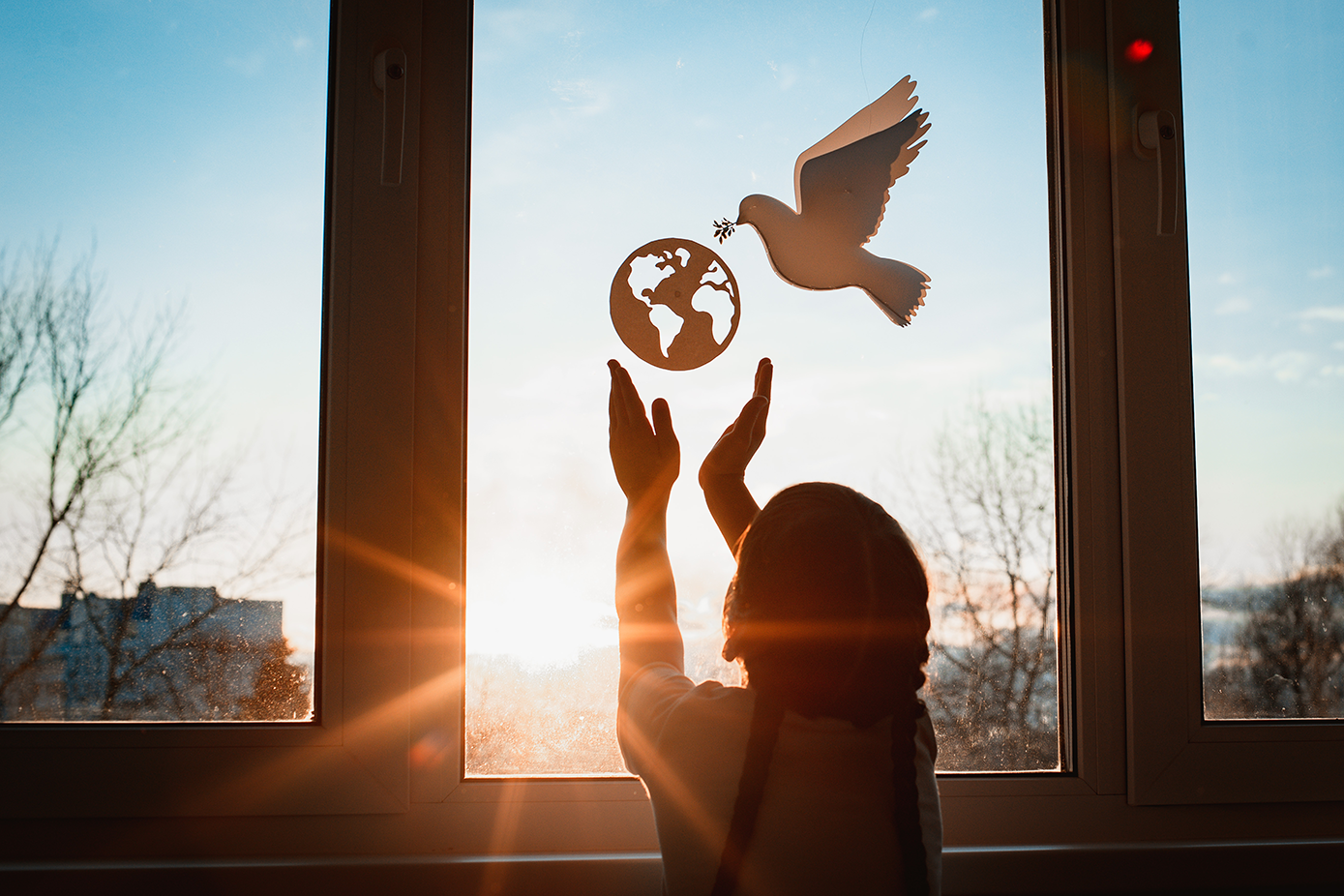 Silhouette of child with outstretched hands reaching toward a cutout of the globe and a dove of peace hanging in a sunlit window at dusk or dawn