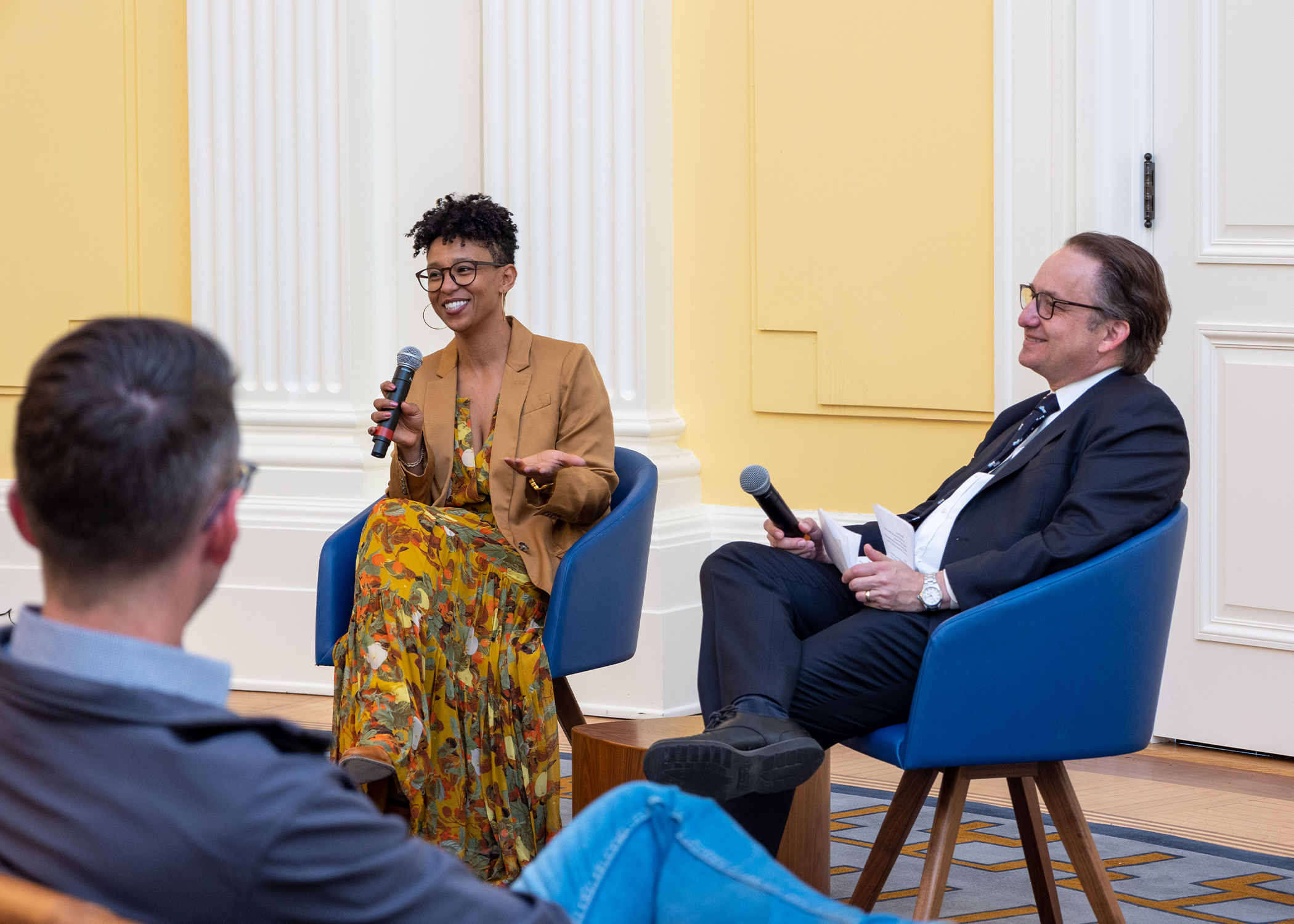 Two people sit in front of an audience. They are speaking to each other using microphones.