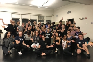 Large group of dance students gathered in a dance studio, smiling at the camera and doing jazz hands