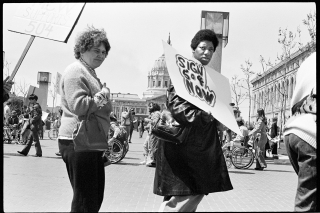 Black and white photograph of protesters with signs marching in a circle in front of federal buildings in San Francisco, CA, 1977.