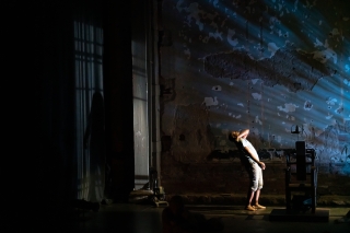 A man, who is bending slightly backwards, is standing on a darkened stage. To his left is a paint chipped-cracked wall.