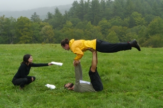 On a green landscape, two people pose in two-person superman yoga pose while a third person to their left holds out a script