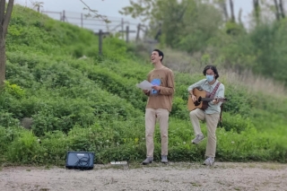 A guitarist and singer perform on a dirt path with a green hill in the background