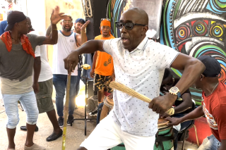 A person in a white shirt and shorts dancing while accompanied by drummers