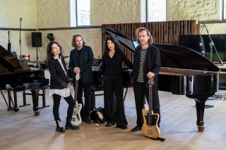 All four members of Dream House Quartet standing in front of two grand pianos. David and Bryce are standing with guitars. A dog is lying on the floor between David and Katia.