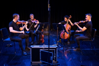 Four members of JACK Quartet with instruments in jeans and a black backdrop.