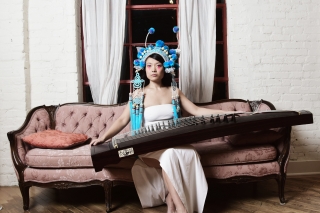 Wu Fei on a Victorian couch, holding an instrument wearing a turquoise headdress..