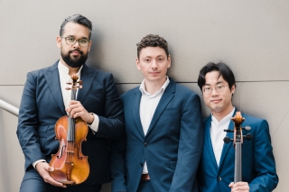 Pianist Dominic Cheli is standing, flanked by Vijay Gupta standing on the left holding a cello and Yoshika Masuda standing on the right holding a cello. All three are wearing matching blue suits.