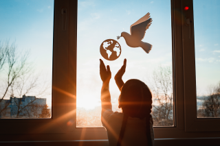 Silhouette of child with outstretched hands reaching toward a cutout of the globe and a dove of peace hanging in a sunlit window at dusk or dawn