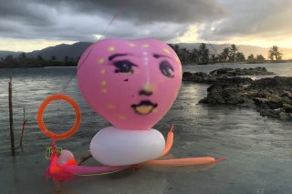 Pink heart shaped balloon with face drawn on, in tropical beach environment.