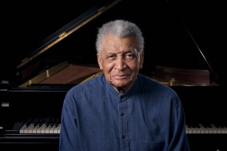 Man stares straight into camera. He is greying and has short cropped hair. He wear a blue linen shirt, and appears in front of a piano.