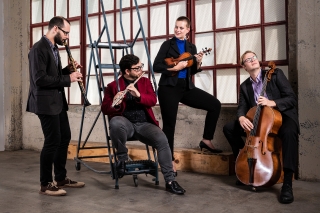 Band in front of tall windows. Playing instruments and smiling.