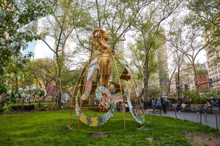 An outdoor sculpture with a golden surface. An open framework resembling a hoop skirt rises from the ground to the waist of a female figure, suspending the figure above the ground.