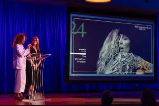 Two women on stage, screen behind them shows image of performance.