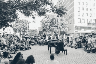 Black and white image of Ibrahim playing a piano outdoors with a crowd. 