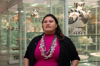 Royce K. Young Wolf, PhD in pink shirt and abundant necklaces stands in front of display cases of artworks.