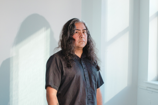 Raven Chacon standing in a white room in front of a small audio mixer and headphones on a white table