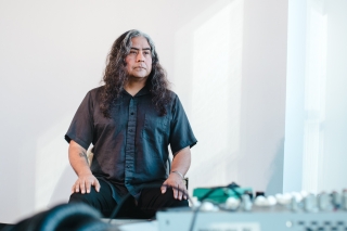 Raven Chacon standing in a room with a blurred mixer and headphones on a table in the foreground