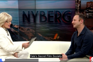 Two people sit together on a cream colored couch in a TV studio. 