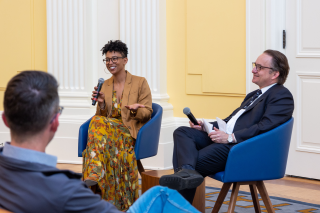 Two people sit in front of an audience. They are speaking to each other using microphones.