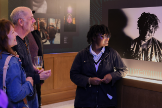 Two people admire student artwork. The artist is next to their photograph.