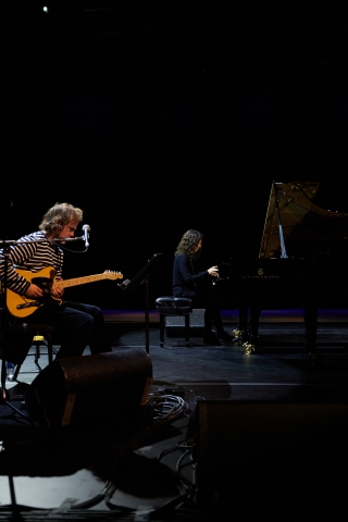 Bryce Dessner seated in the foreground performing on guitar while Katia Lebeque performs piano in the background