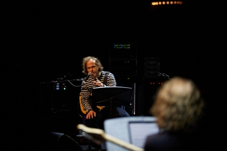 Bryrce Dessner speaking into a microphone while seated on stage holding a guitar in his lap