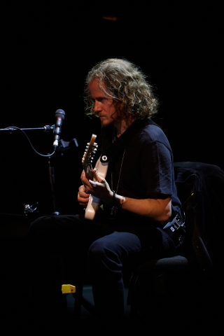 David Chalmin sitting in a chair performing guitar on stage