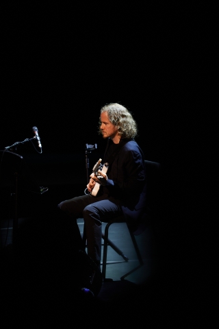 David Chalmin sitting performing on a chair preforming guitar on stage