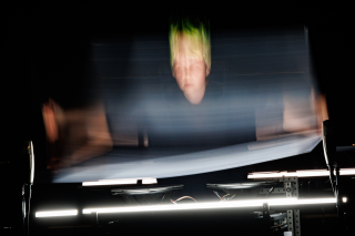 A motion blurred image of Ash Fure shaking a plexiglass board, arms outstretched