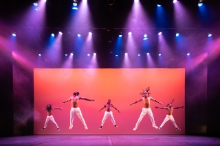 Five dancers jumping in the air, on orange stage with purple lighting.