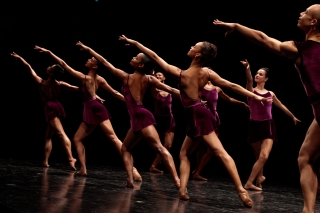 A row of dancers in directed lighting in a reaching pose