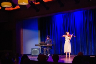 Woman playing a violin, on stage, with a keyboardist. 