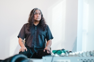 Raven Chacon standing in a room with a blurred mixer and headphones on a table in the foreground